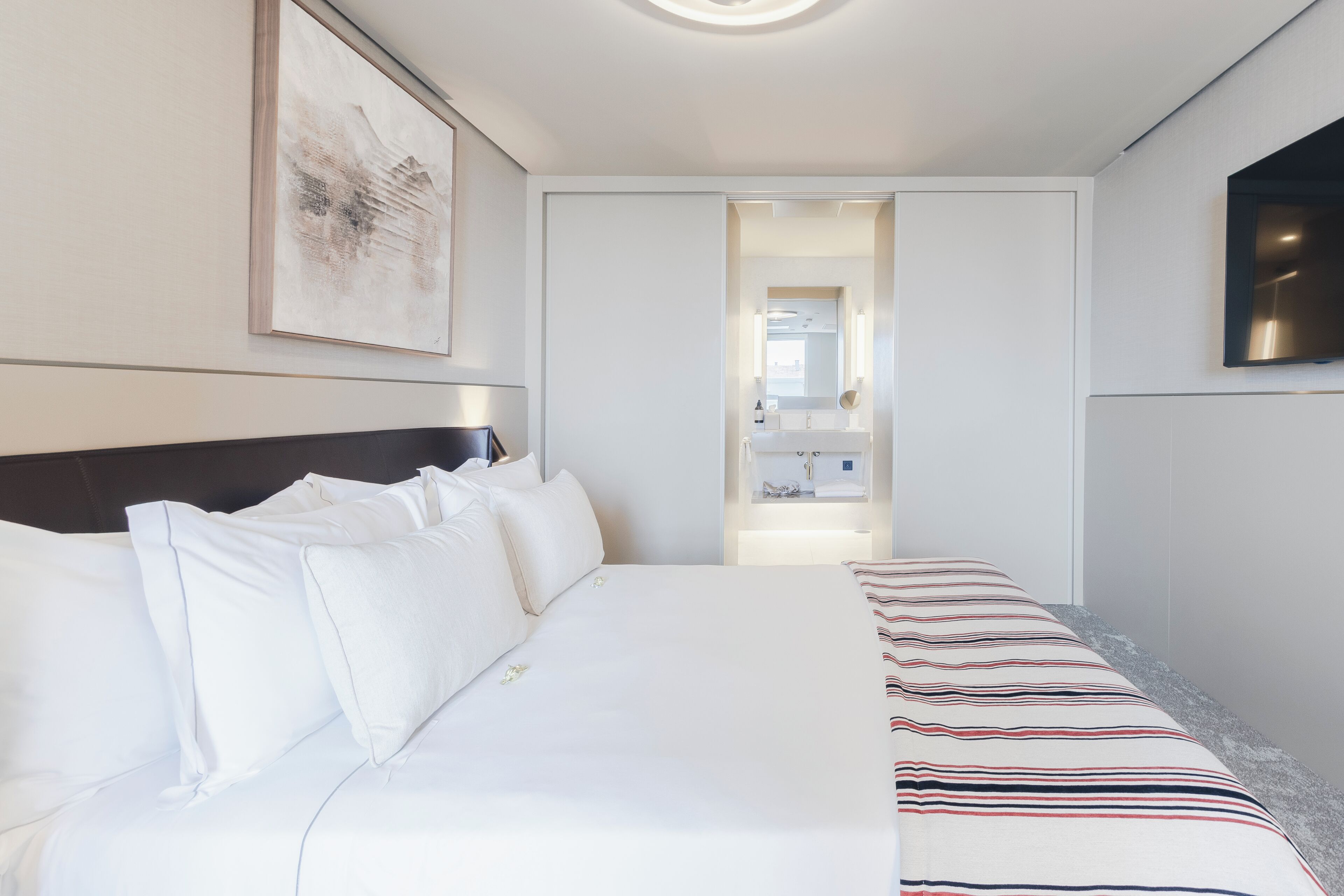 A hotel room with a bed, a flat-screen TV, a framed artwork, and a bathroom visible through an open door.