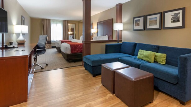 A hotel room with a bed, a blue sofa, a brown ottoman, a desk with a lamp, and a TV. - Comfort Suites Billings (Billings)