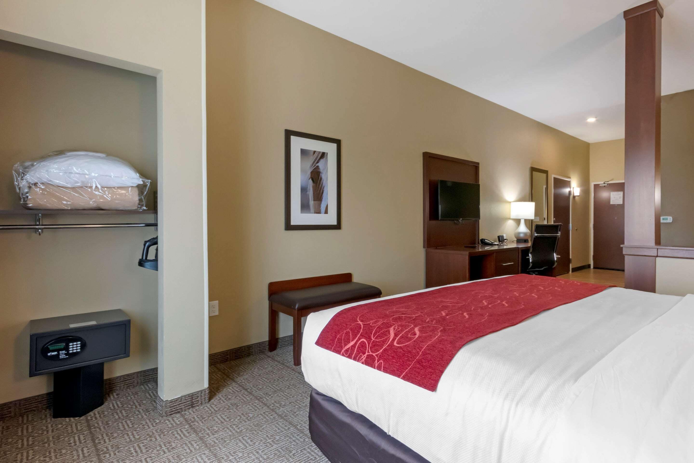 A hotel room with a bed, a desk, a chair, a small table, a safe, a mirror, a television, and a wall-mounted shelf with a pillow.