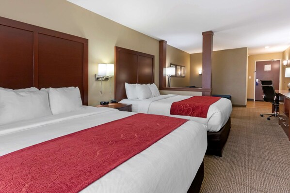 Ein Hotelzimmer mit zwei Betten, jeweils mit weißer Bettwäsche und einer roten Tagesdecke. Es gibt einen Schreibtisch mit einem Stuhl, eine Lampe und einen Spiegel an der Wand. Der Raum hat einen Teppichboden und ein hölzernes Kopfteil.