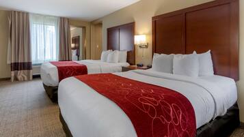 A hotel room with two beds, each with white linens and red throw blankets. There is a wooden headboard, a bedside table with a lamp, and a large window with curtains.