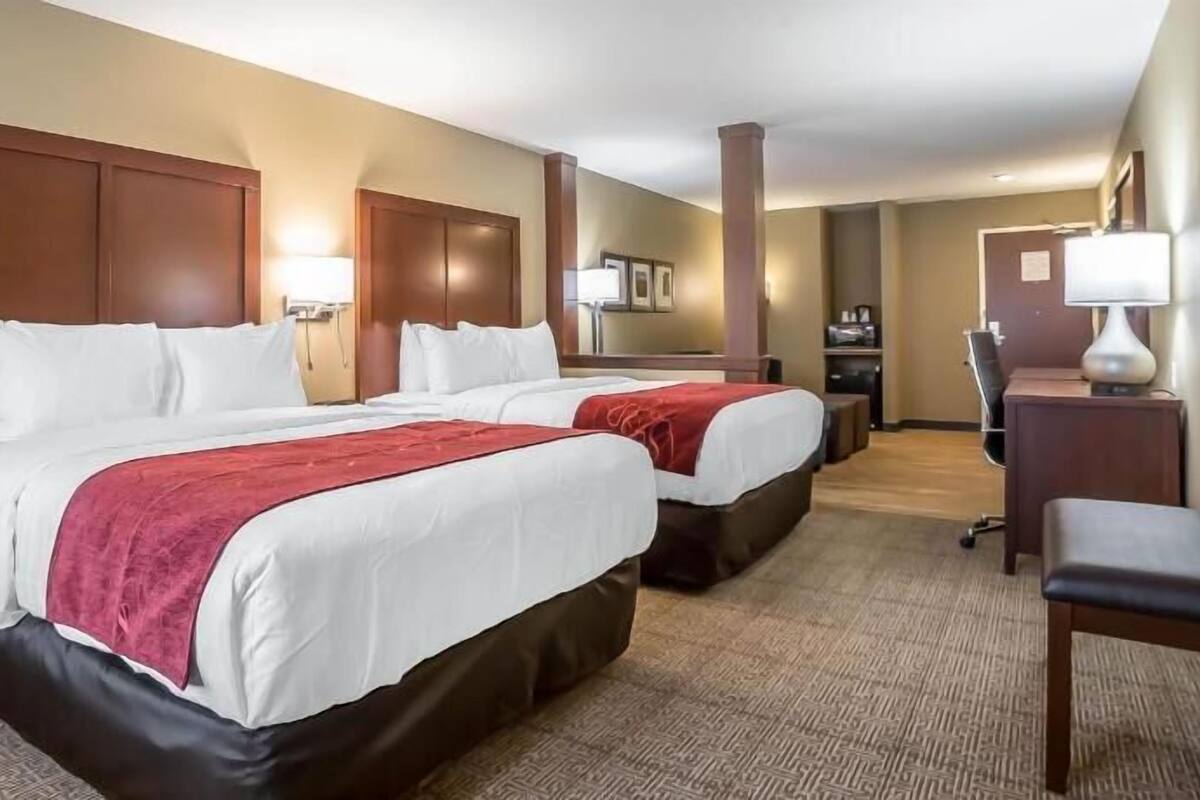 A hotel room with two beds, a desk, a chair, and a lamp.