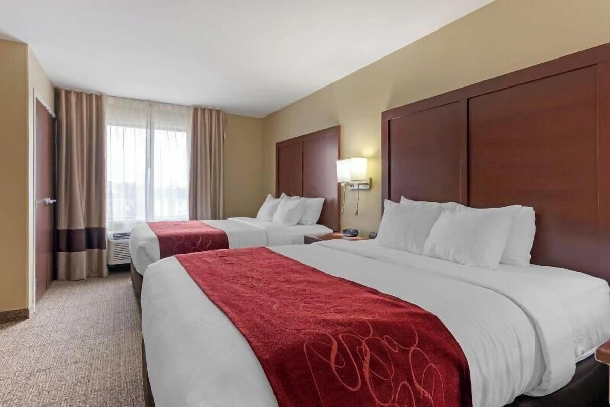 A hotel room with two beds, a large window with curtains, a wall-mounted lamp, and a wooden headboard.