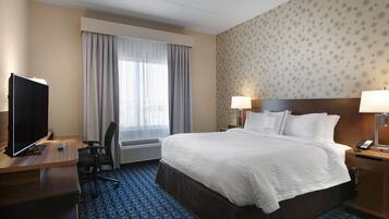 A hotel room with a bed, a desk with a TV, a chair, a window with curtains, and a patterned wall.