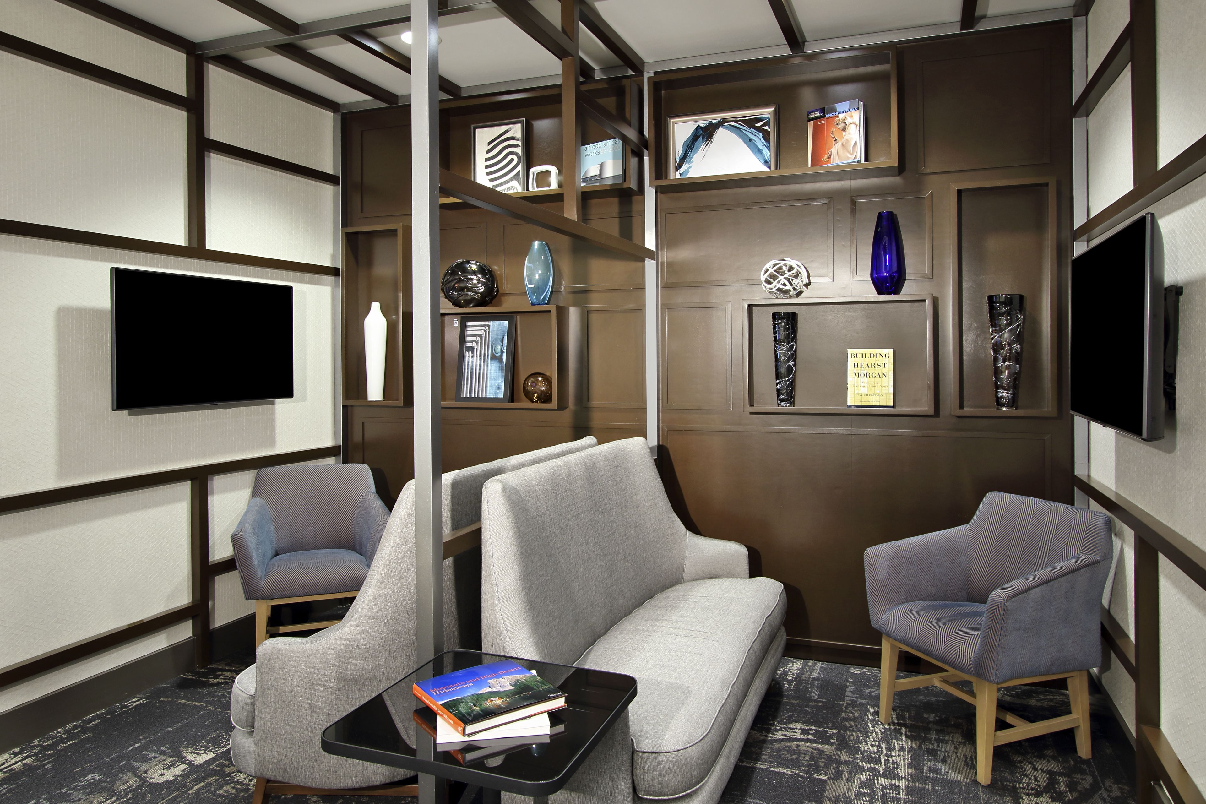 Een moderne hotelkamer met een grijze bank, twee fauteuils, een flatscreen televisie, een boekenkast met decoratieve objecten en een zwarte salontafel met boeken.