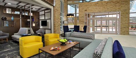 A modern lobby with a grey sofa, yellow armchairs, a wooden coffee table with fruit, and a large window offering a view of the outside.