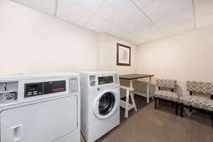 A laundry room with two commercial dryers, a table, and two patterned chairs.