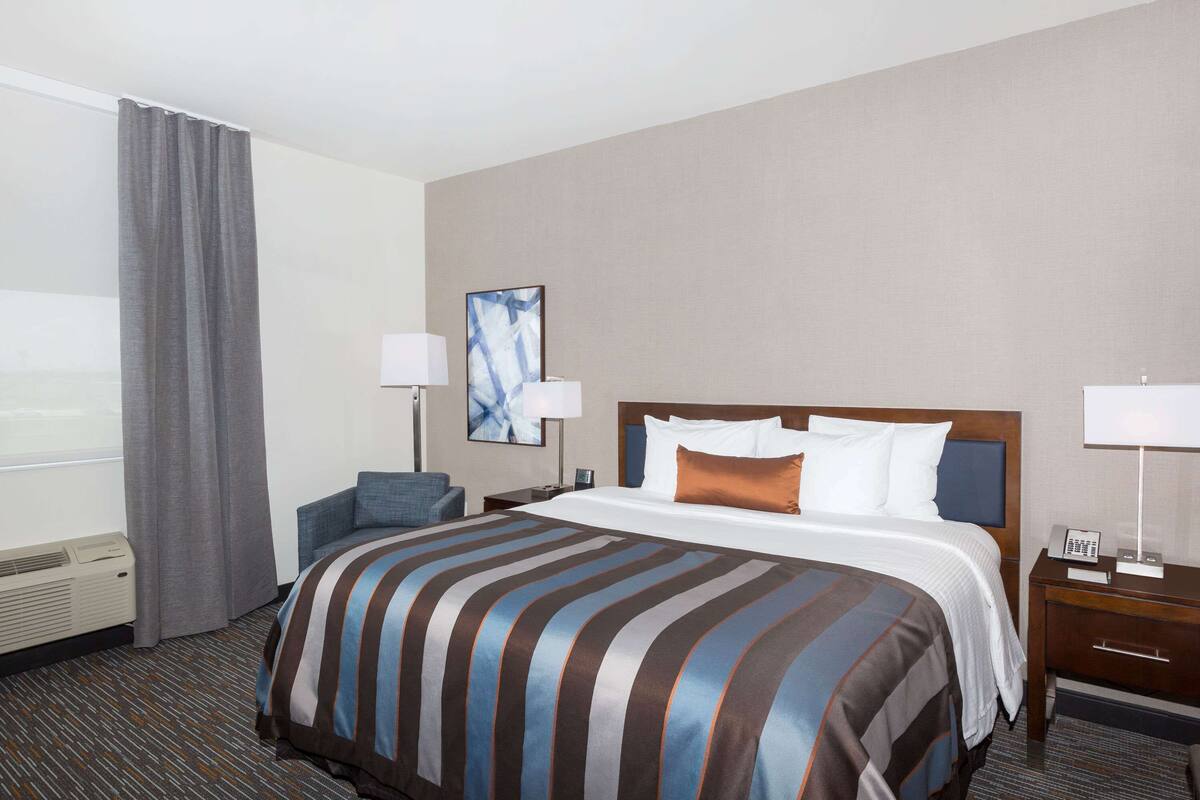 A hotel room with a large bed, a chair, a desk with a lamp, and a painting on the wall.