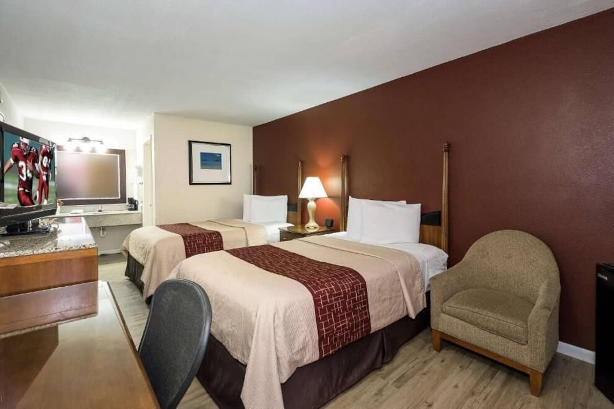 A hotel room with two beds, a desk, a chair, a TV, and a lamp.