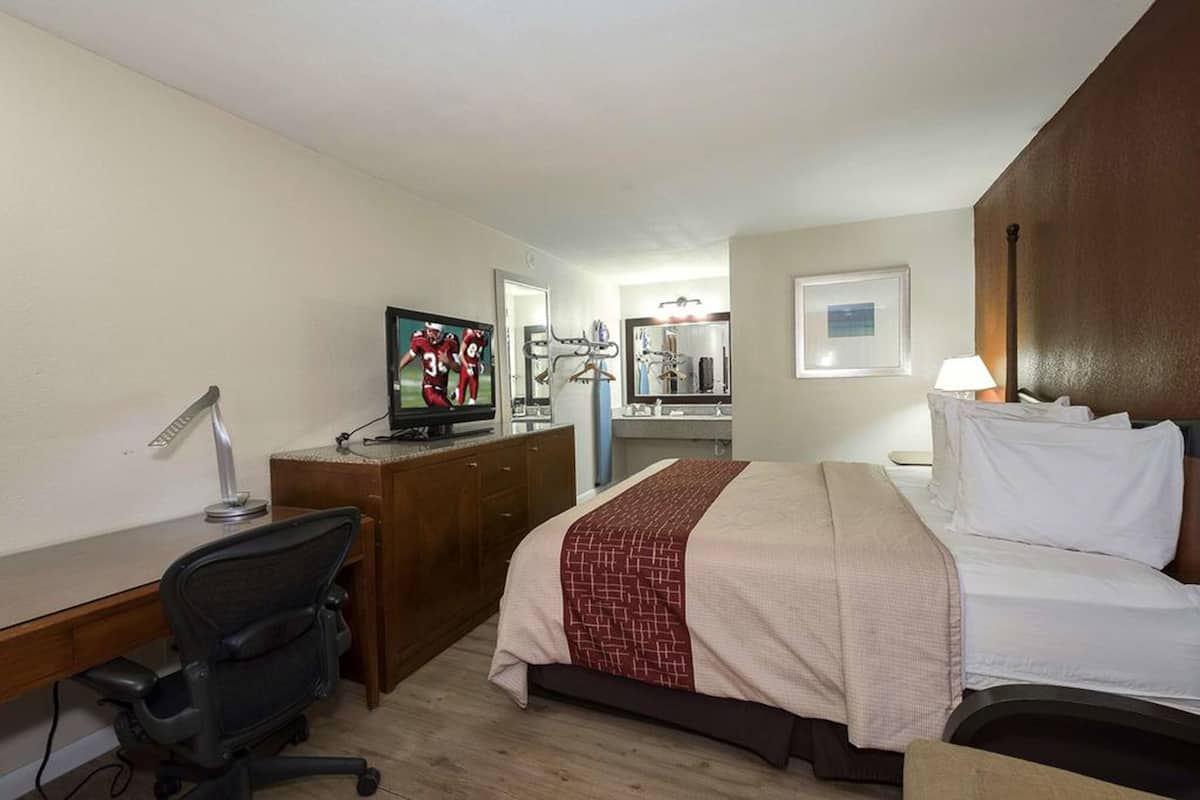 A hotel room with a bed, desk, chair, TV, and a bathroom with a mirror and sink.