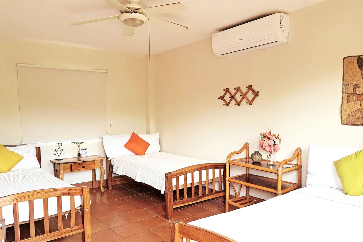 A room with two single beds, a wooden table with chairs, a ceiling fan, and an air conditioning unit.