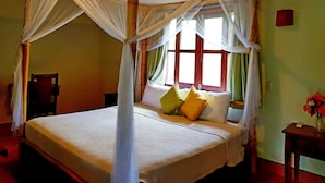 A bed with a canopy, a chair, a bedside table with a vase of flowers, and a window with curtains. - Hotel Popoyo (Tola)
