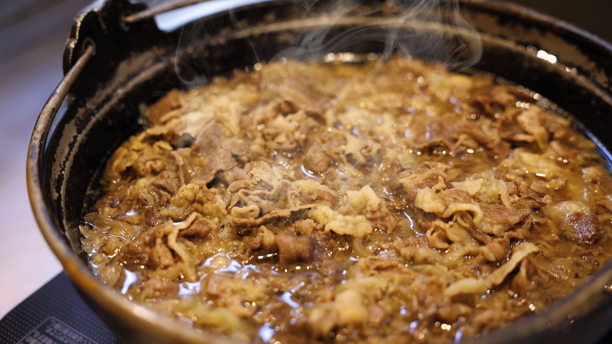 Lähikuva keittoastiasta, jossa on kansi. Astiassa on kuplivaa, höyryävää ruokaa, ja siinä on näkyvissä lihapaloja ja kasviksia.