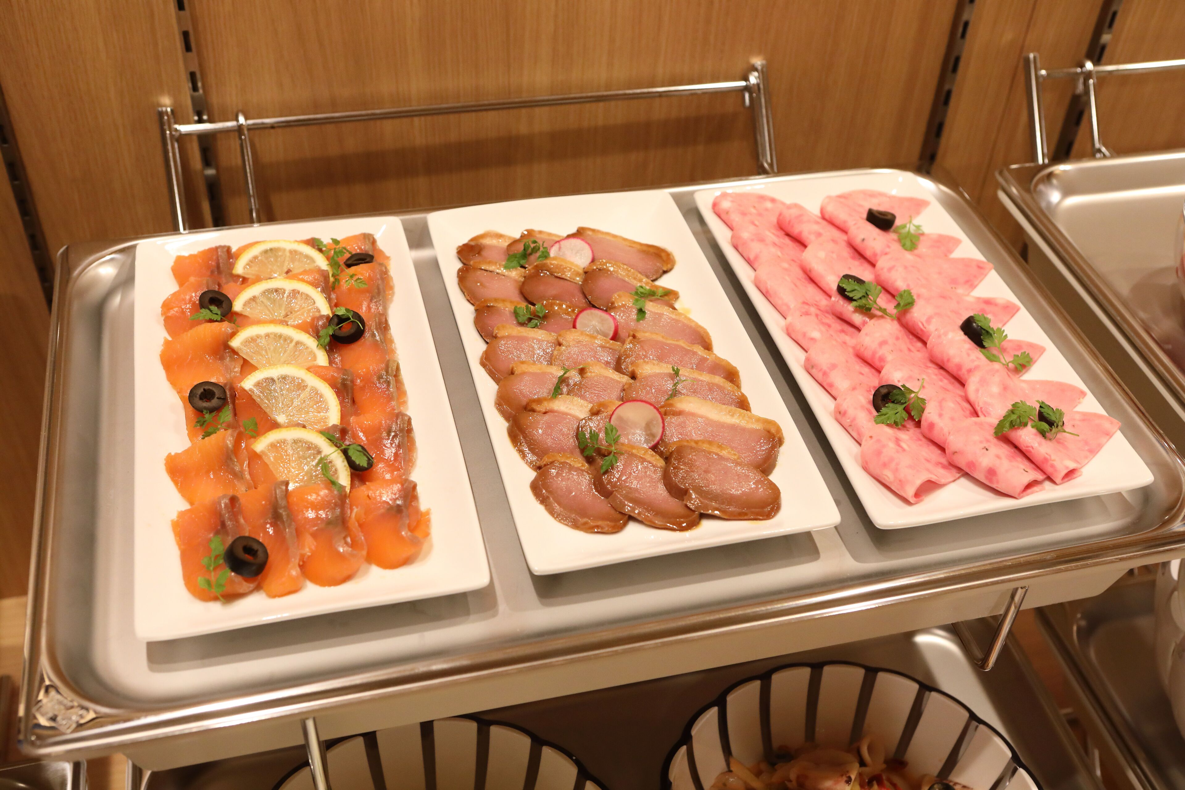 Una mesa de buffet con tres fuentes de comida: salmón ahumado con rodajas de limón y aceitunas negras, jamón en rodajas con rábanos y perejil, y carne enrollada con hierbas.