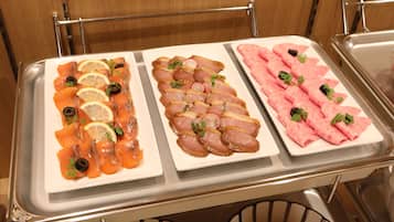 Una mesa de buffet con tres fuentes de comida: salmón ahumado con rodajas de limón y aceitunas negras, jamón en rodajas con rábanos y perejil, y carne enrollada con hierbas.