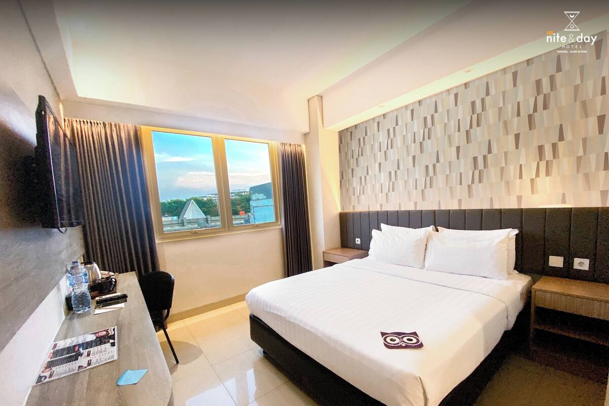 Ein Hotelzimmer mit einem großen Bett, einem flachbildfernseher an der Wand, einem Schreibtisch mit Stuhl und einem fenster mit Vorhängen.