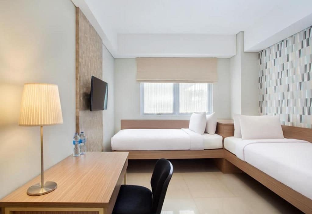 Habitación de hotel con cama, escritorio, silla, televisor y ventana con persianas.