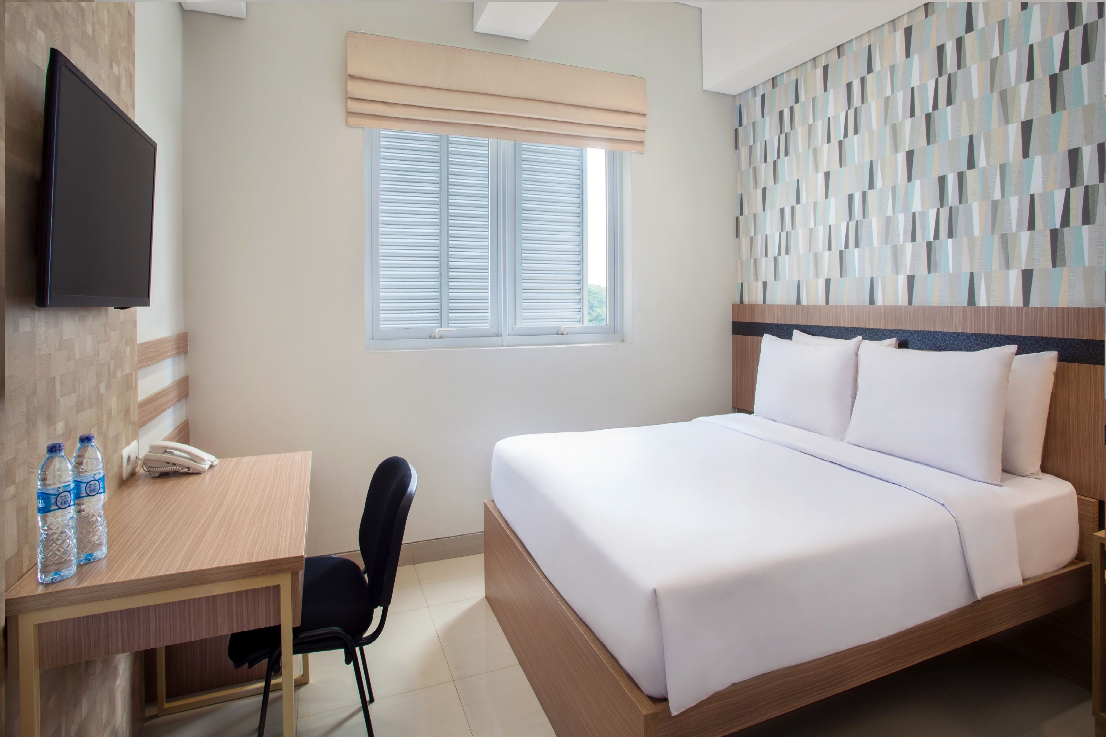 A hotel room with a bed, a desk with a chair, a TV, and a window with blinds.