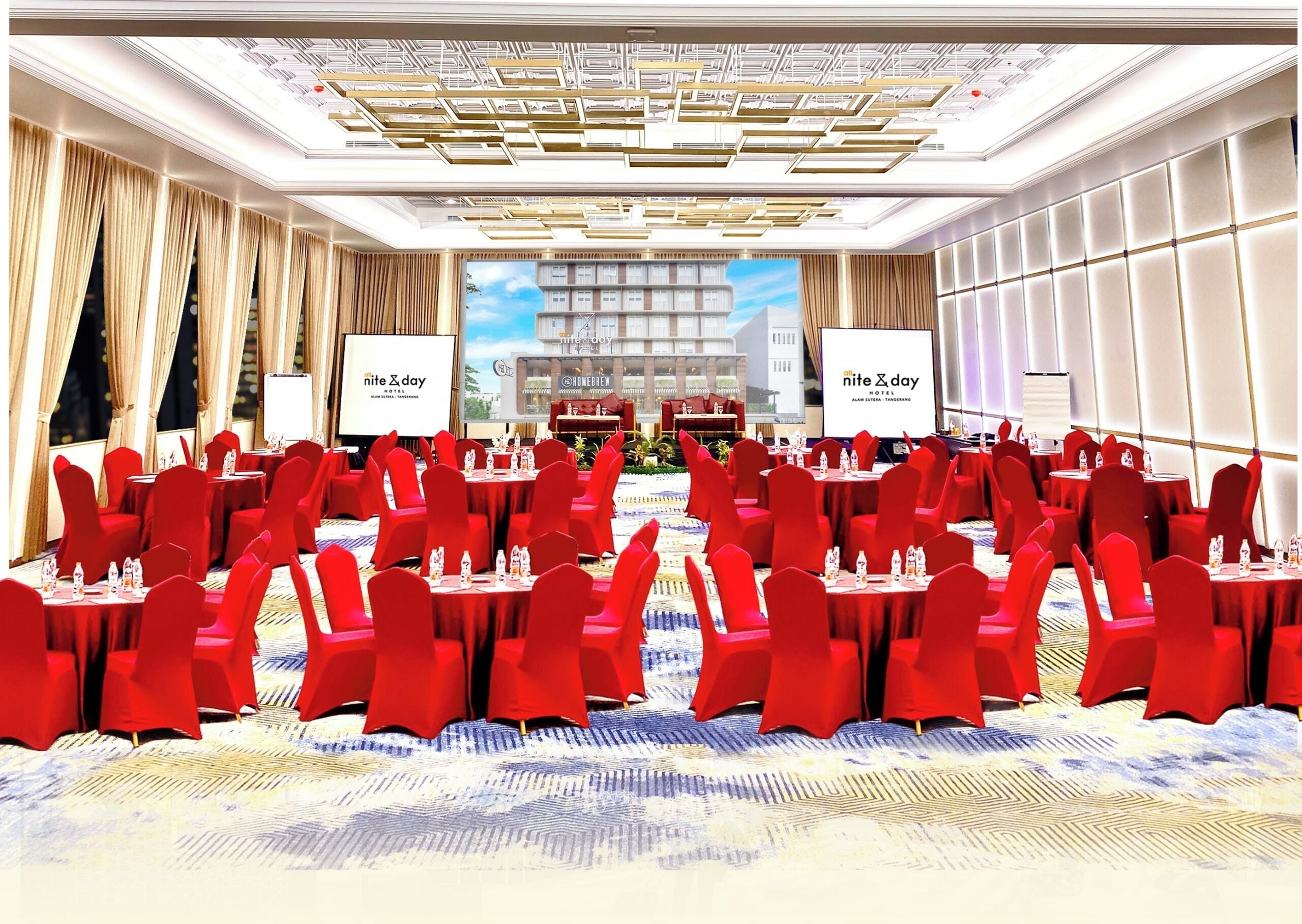 Un salón de conferencias con sillas y mesas rojas, alfombra estampada y una pantalla grande mostrando el nombre del hotel.