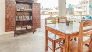 Ein Bücherregal aus Holz mit Büchern, ein Marmortisch und Stühle in einem Raum mit Betonboden und Blick auf Gebäude.