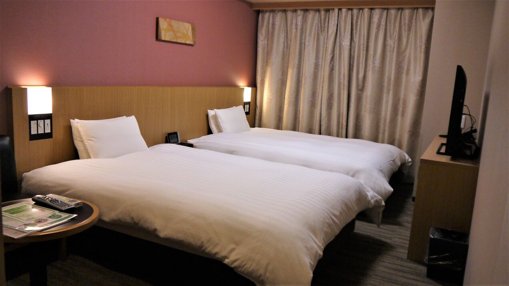 A hotel room with two beds, a nightstand with a remote control and a magazine, a clock, and a television on a stand.