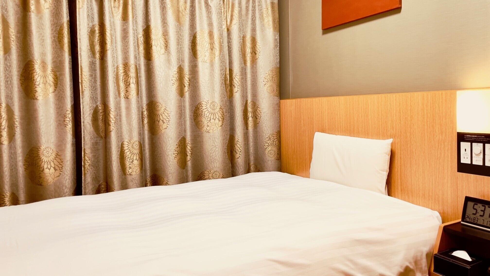 A hotel room with a bed, curtains, a wooden headboard, a nightstand, and a digital clock.
