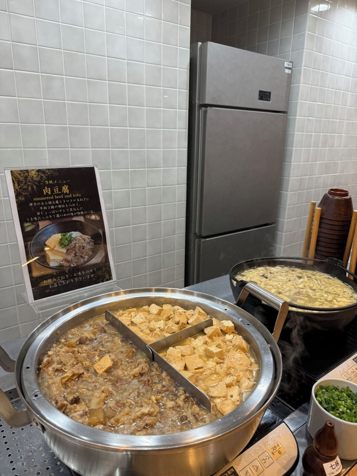 Un buffet proposant du bœuf et du tofu mijotés, une terrine de soupe et un bol d’oignons verts.