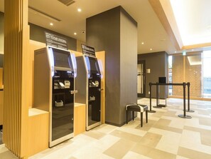 Een moderne hotellobby met zelfbedieningskiosken, een zithoek en een glazen scheidingswand.