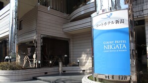 Exterior del Hotel Court Niigata, con fachada de baldosas, un llamativo letrero azul y un área de entrada ajardinada.