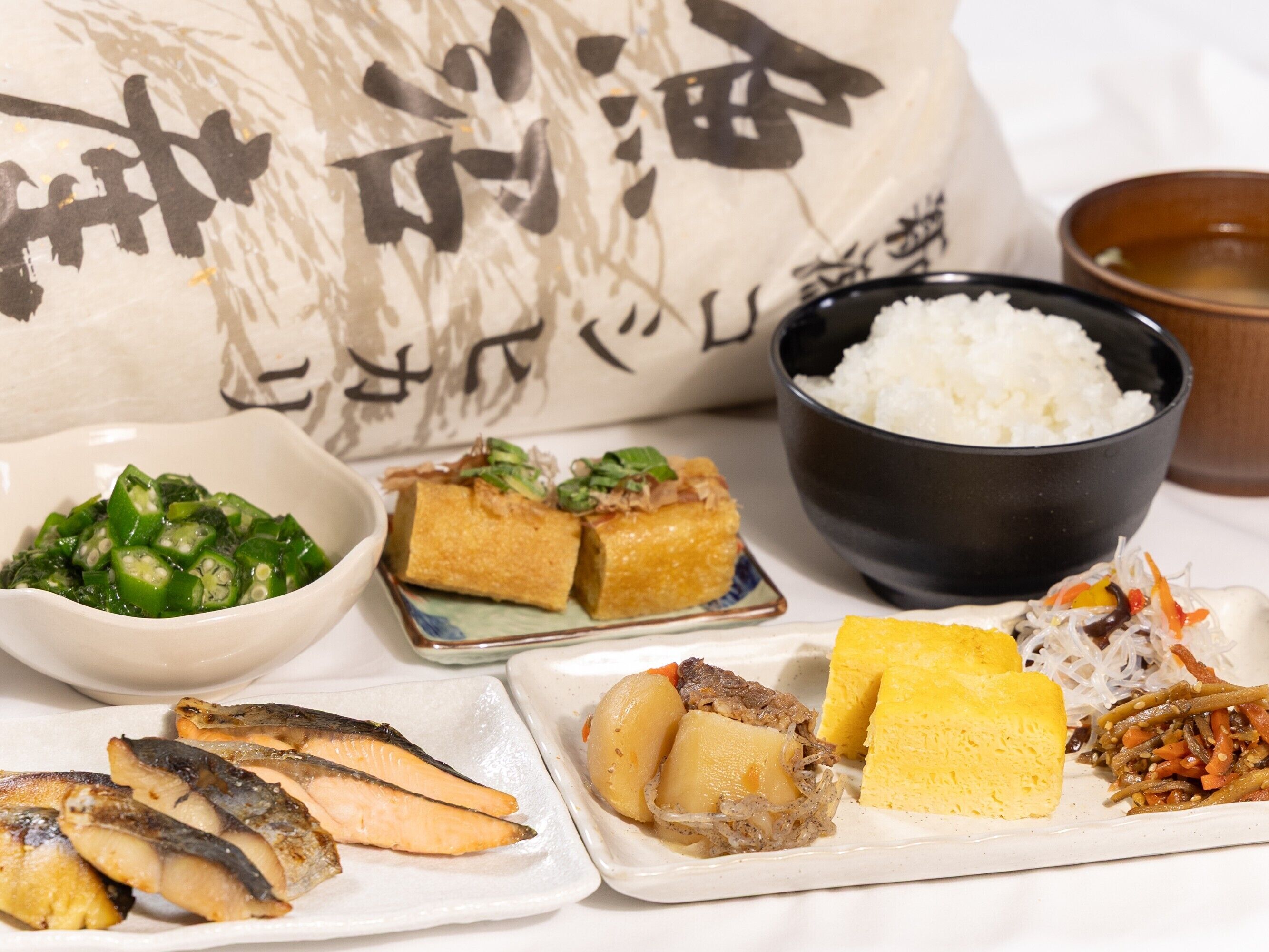 A variety of Japanese dishes including fish, tofu, and vegetables, served on a white table with a black bowl of rice and a decorative pillow in the background.