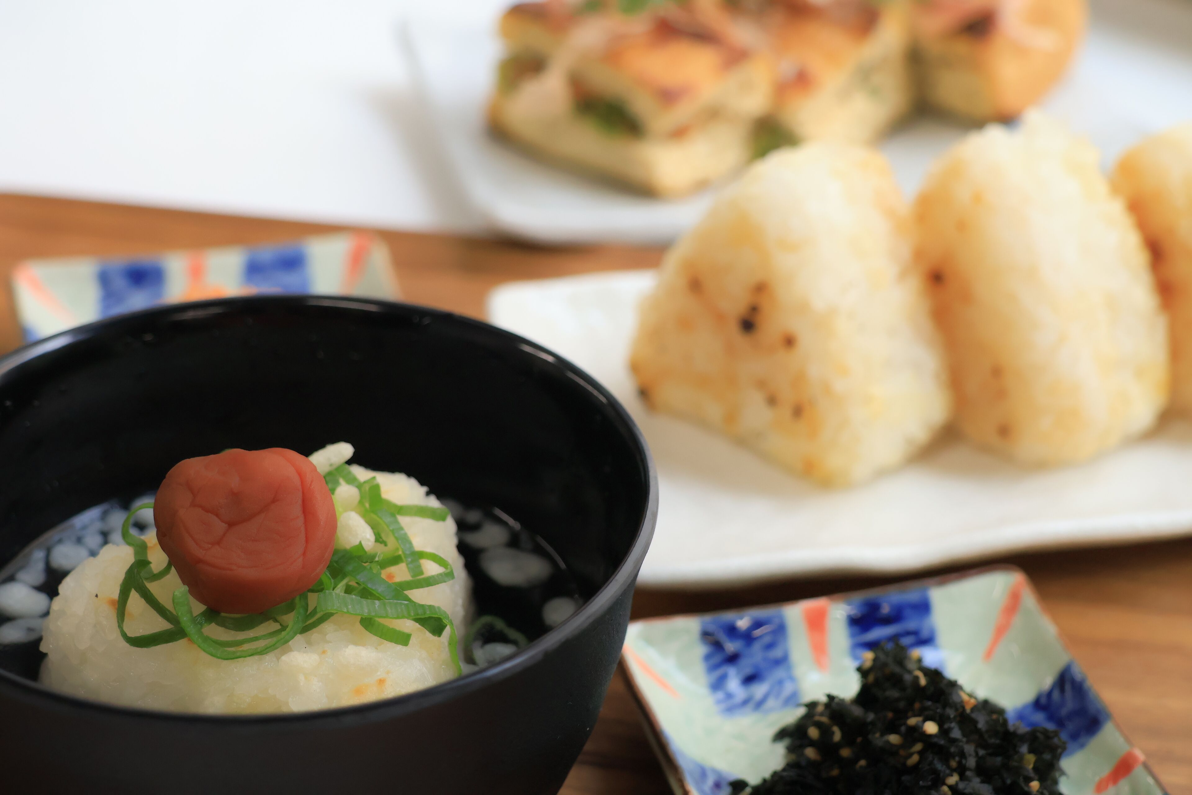 A bowl of rice with a decorative item and green garnish, a plate with three triangular food items, and a small dish of black sesame seeds.