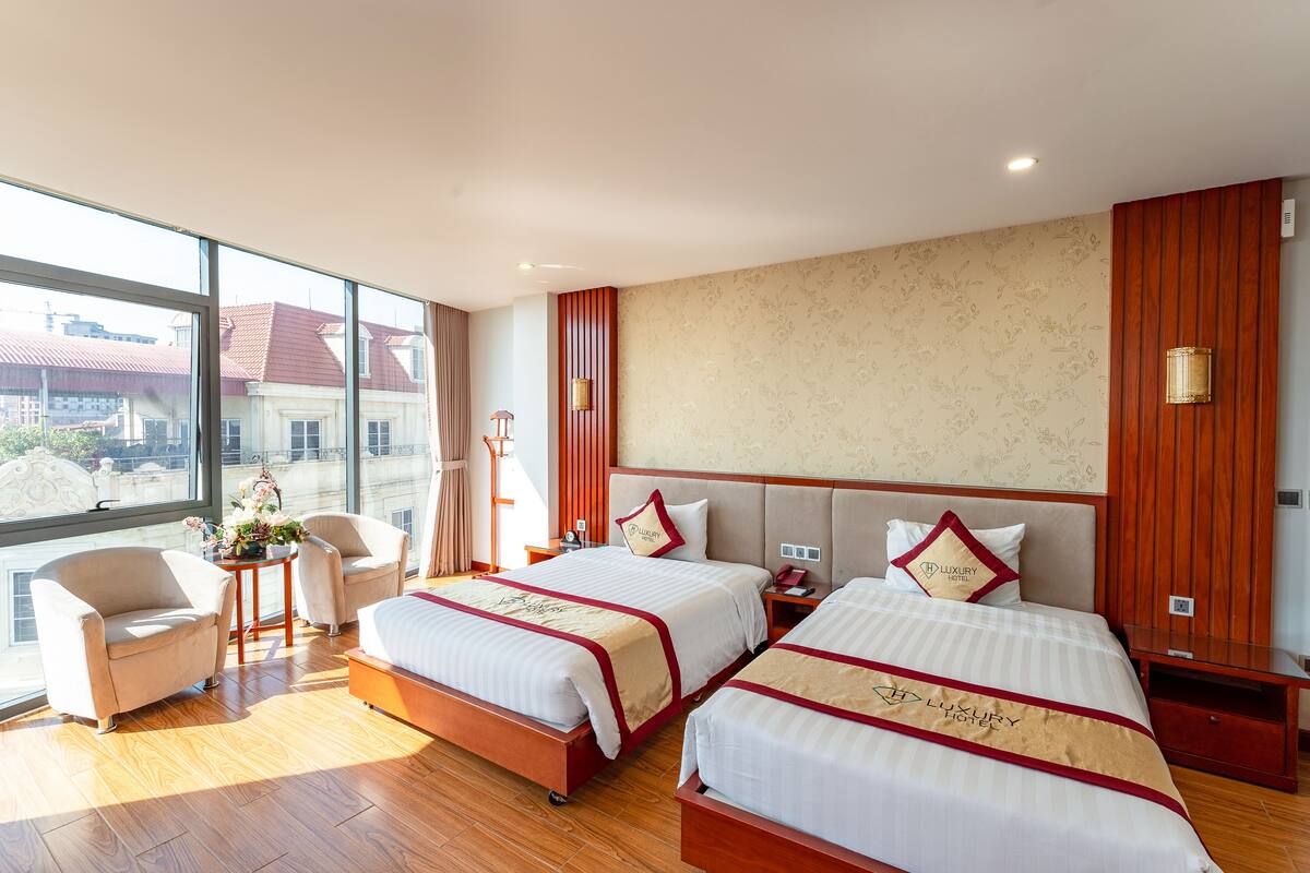 Ein Hotelzimmer mit zwei Betten, einer Sitzecke mit einem Sessel, einem kleinen Tisch mit einer Vase und großen Fenstern mit Blick auf Gebäude.
