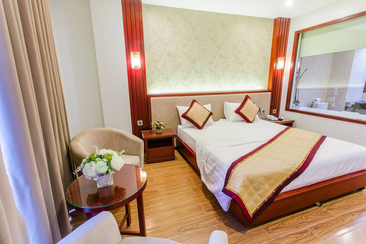 Ein Hotelzimmer mit Bett, Sessel, kleinem Tisch mit Blumenvase und Nachttisch mit Lampe.