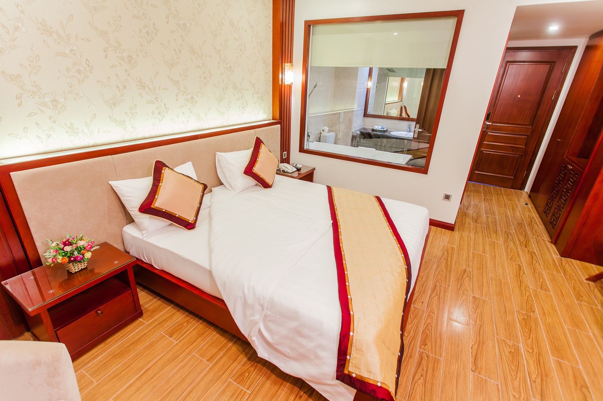 A hotel room with a large bed, a nightstand with a lamp, a small table with a vase of flowers, and a large mirror above the bed.
