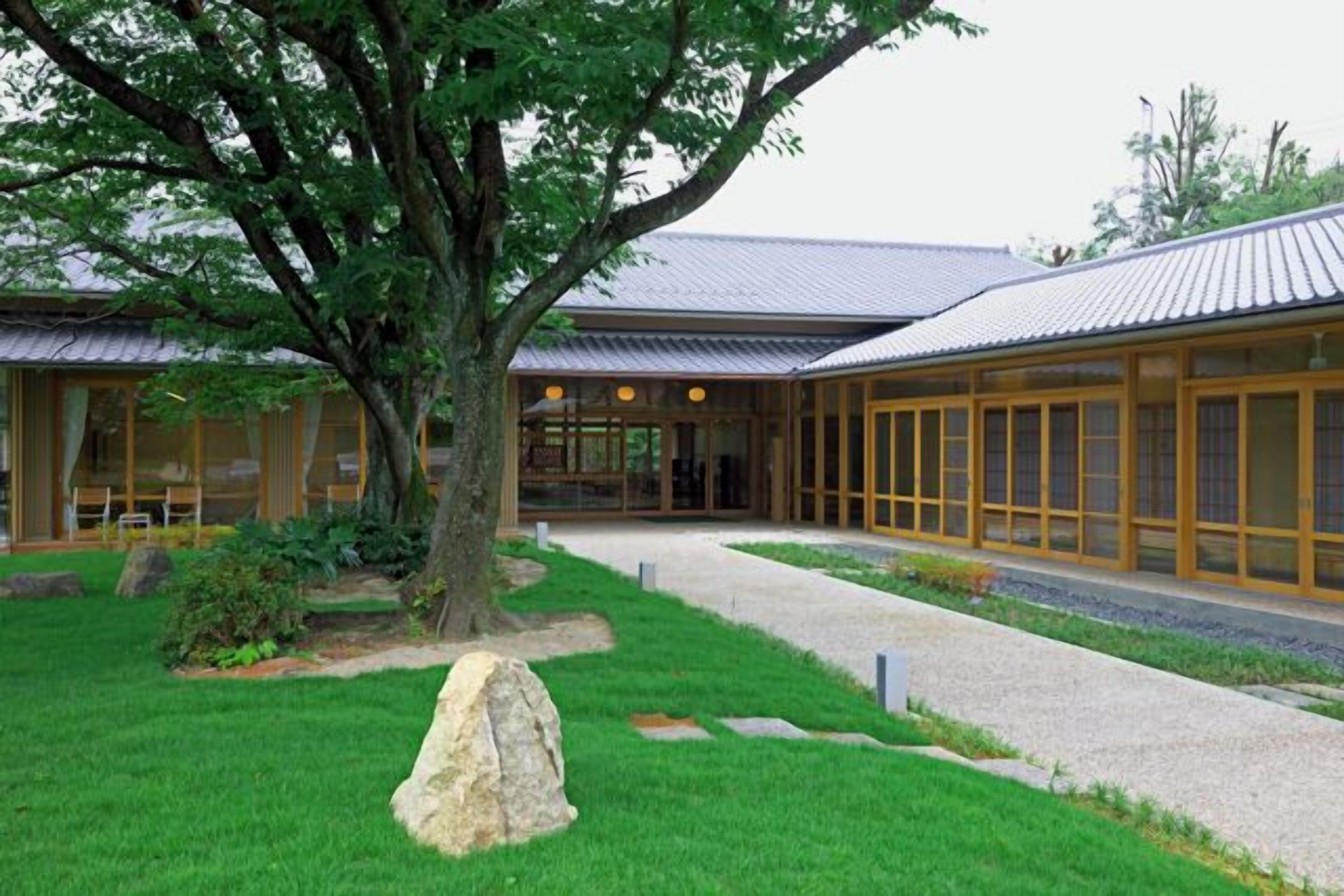 A building with a traditional architectural style, featuring a large tree in the center, a well-maintained lawn, and a pathway leading to the entrance.
