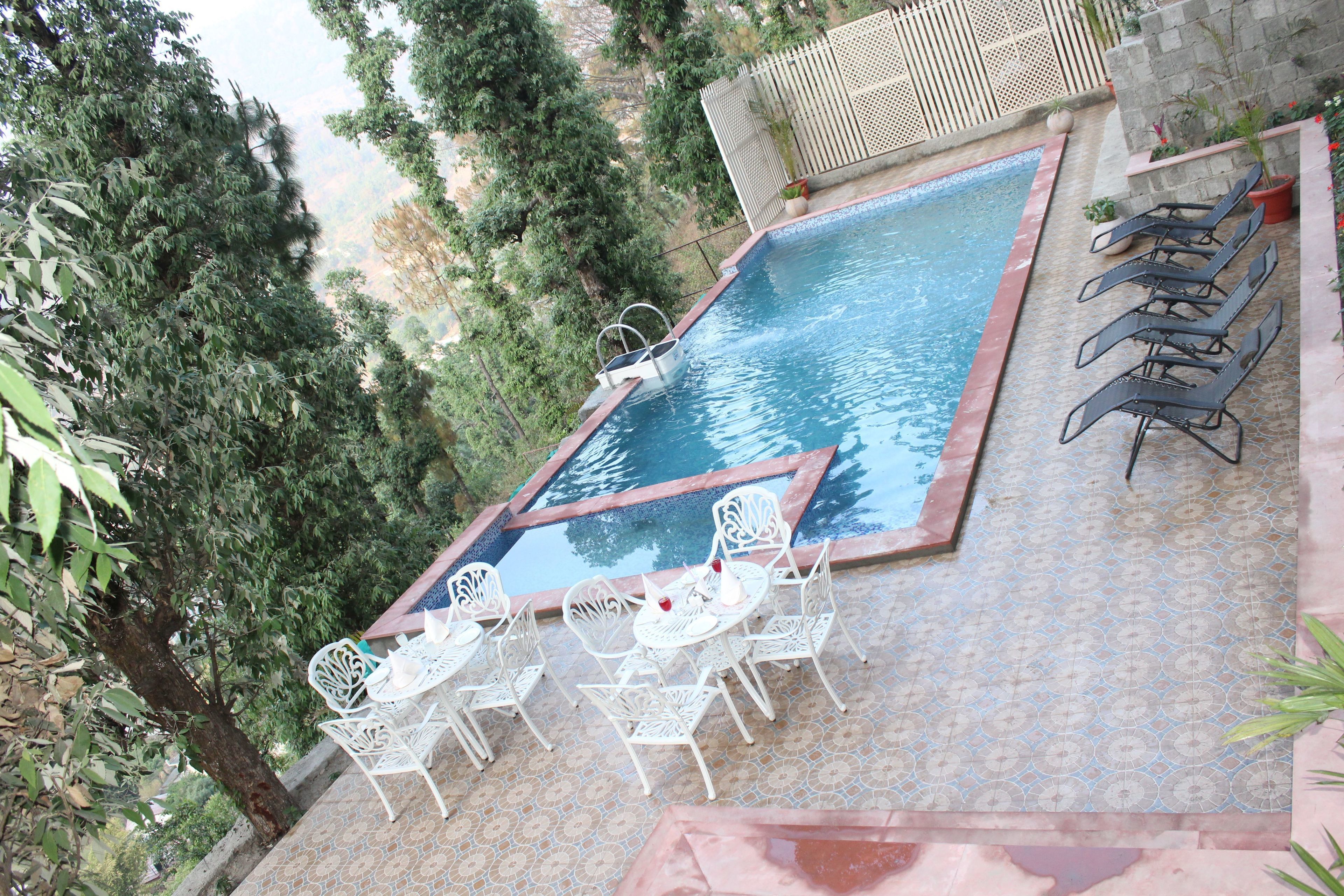 A swimming pool with a small hot tub, surrounded by white metal chairs and tables, set against a backdrop of trees and a stone wall.