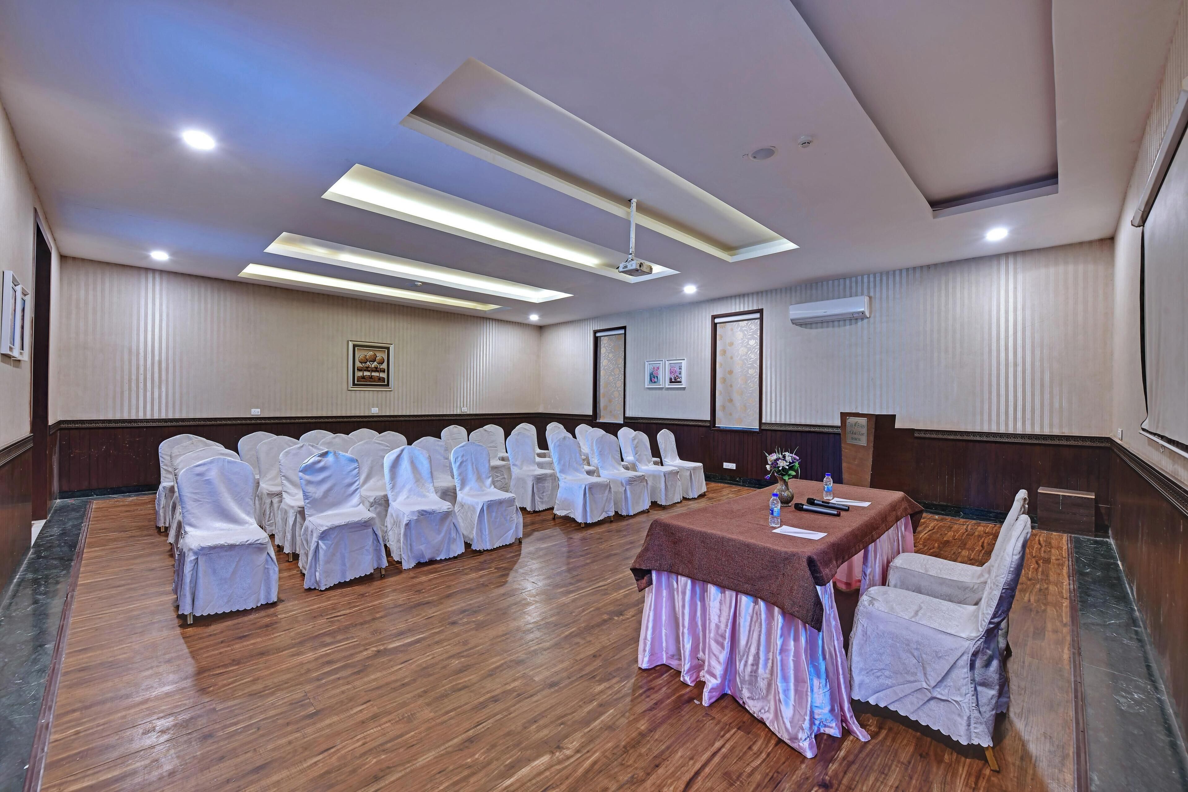 Un salón de conferencias con filas de sillas, una mesa y un proyector en el techo.
