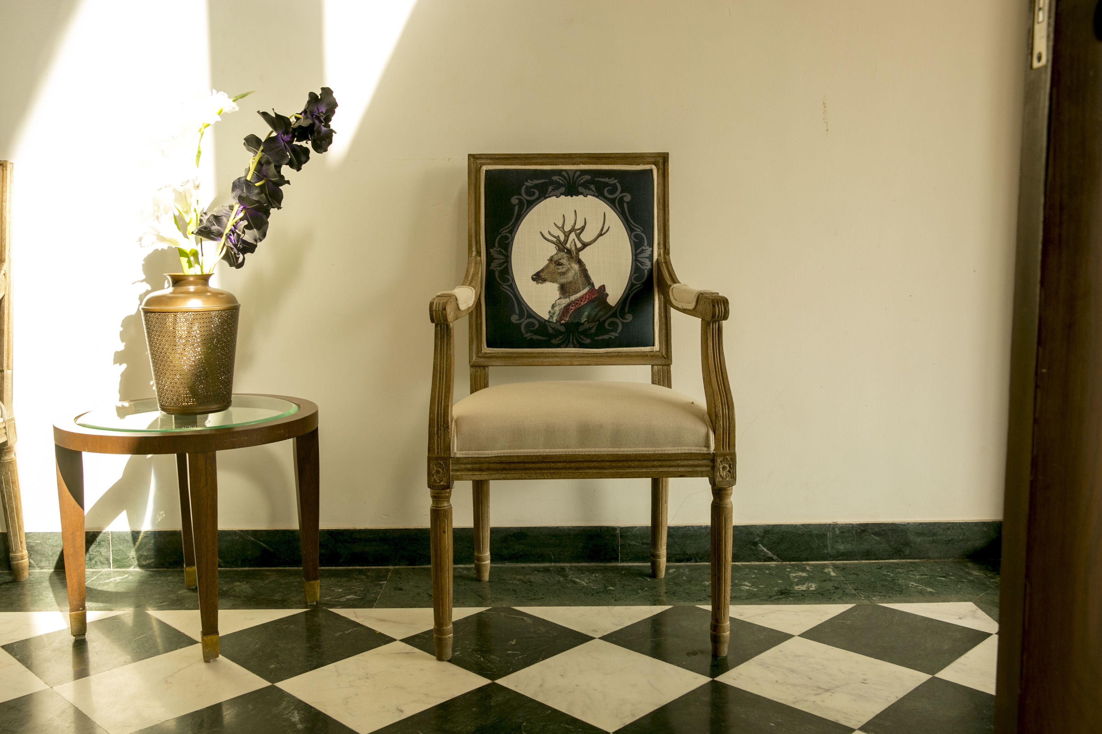 A room with a checkered floor, a chair with a framed picture, and a vase with flowers.