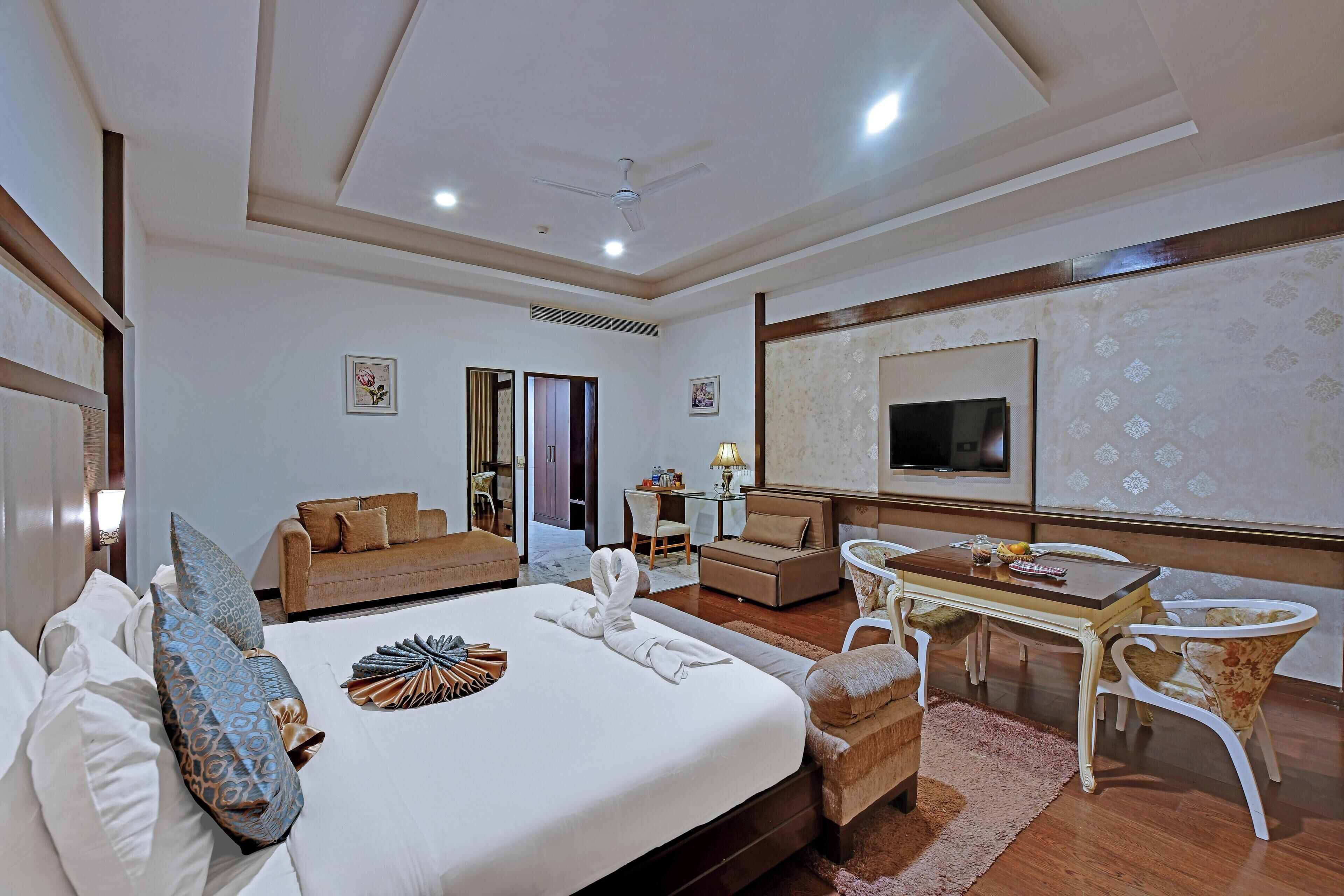 Habitación de hotel con cama, dos sofás, una mesa de comedor, televisor y ventilador de techo.