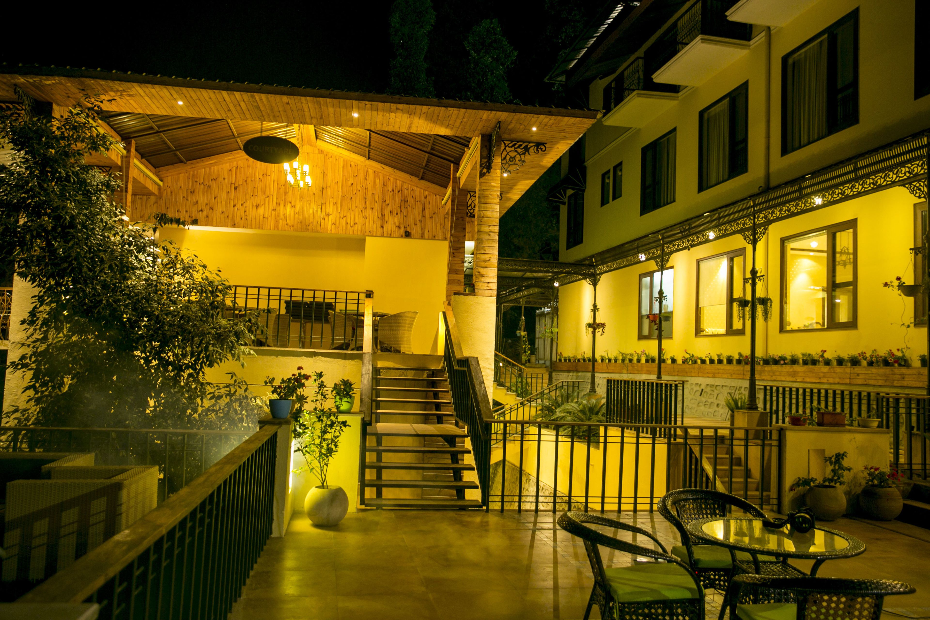 A covered outdoor area with a staircase leading to an upper level, a seating area with chairs and a table, and a building with multiple windows and balconies.