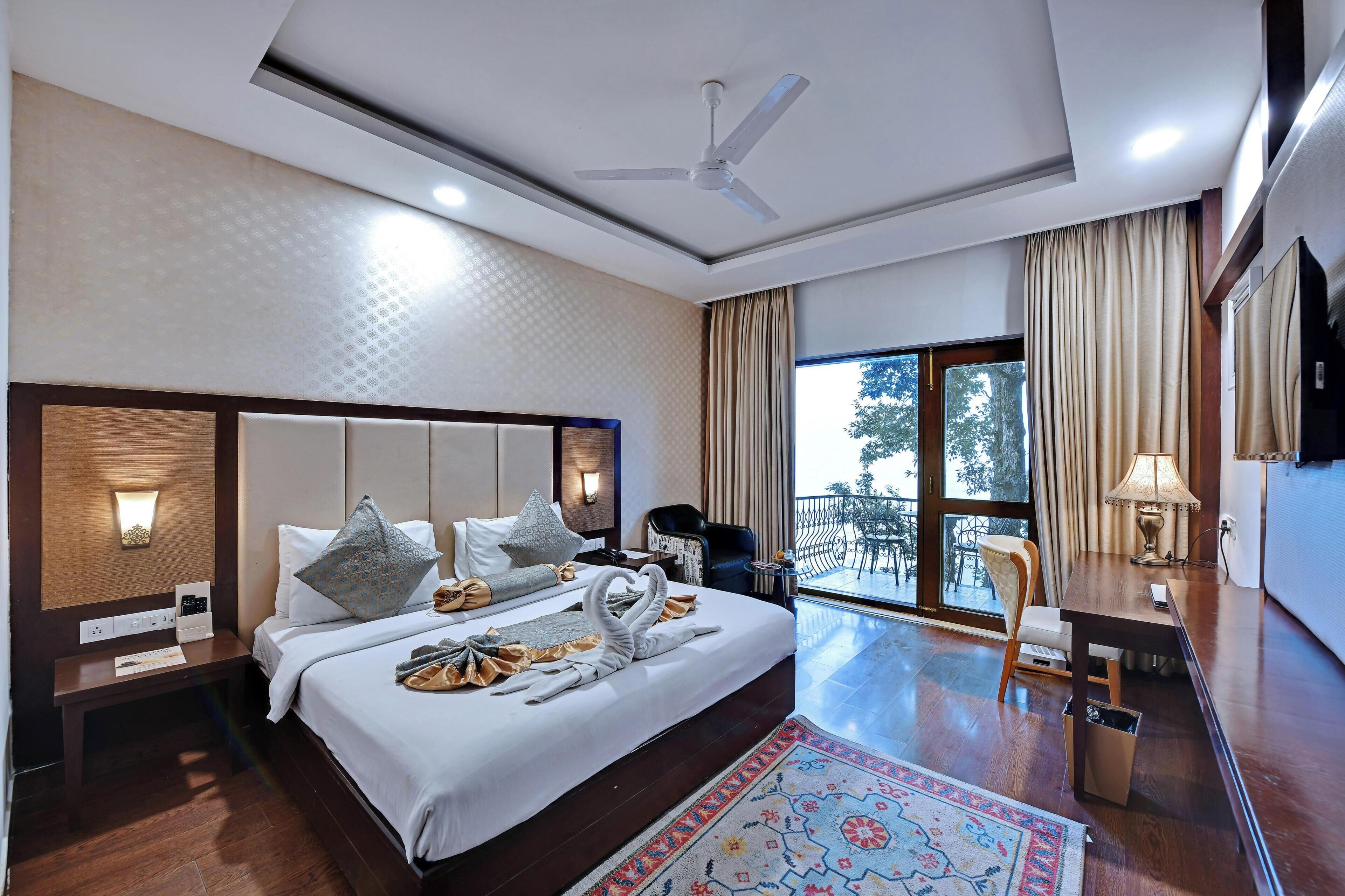 A hotel room with a bed, a desk, a chair, a balcony with a view, and a ceiling fan.