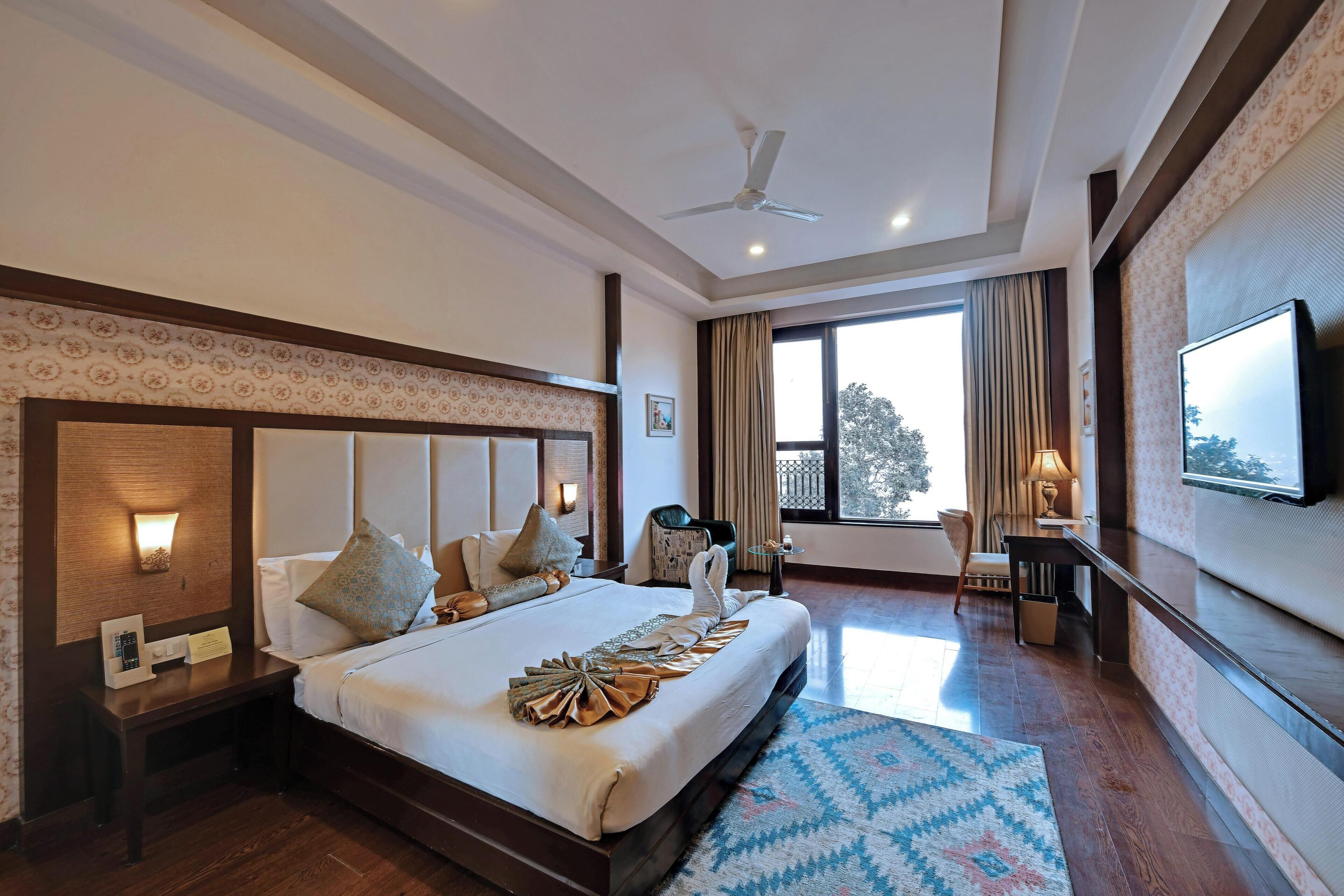 A hotel room with a large bed, a ceiling fan, a TV, and a window with curtains.