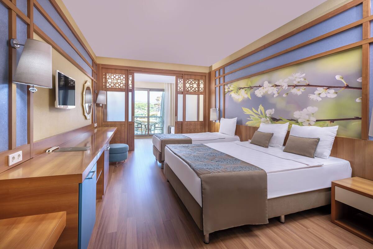 A hotel room with a wooden floor, two beds, a desk with a lamp, a TV, and a decorative wall with a floral design.