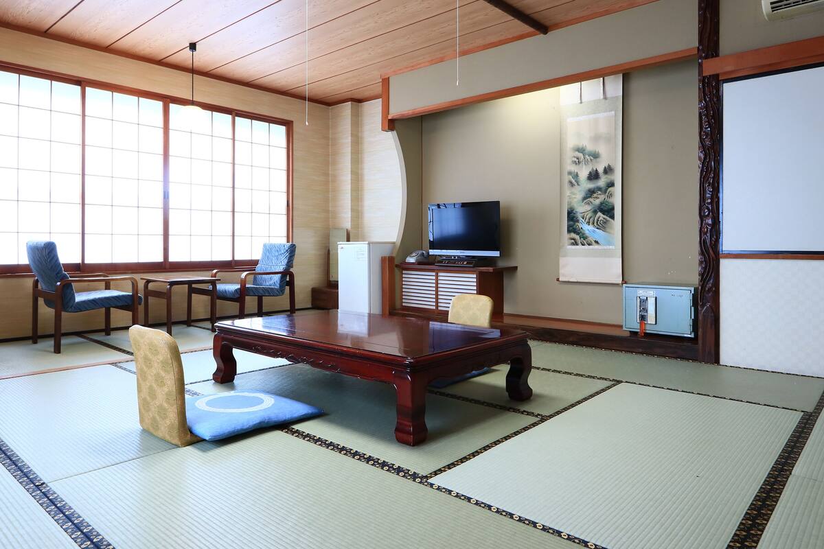 Une pièce de style japonais traditionnel, avec un sol recouvert de tatamis, une basse table en bois, deux chaises bleues, un téléviseur et un tableau accroché au mur.