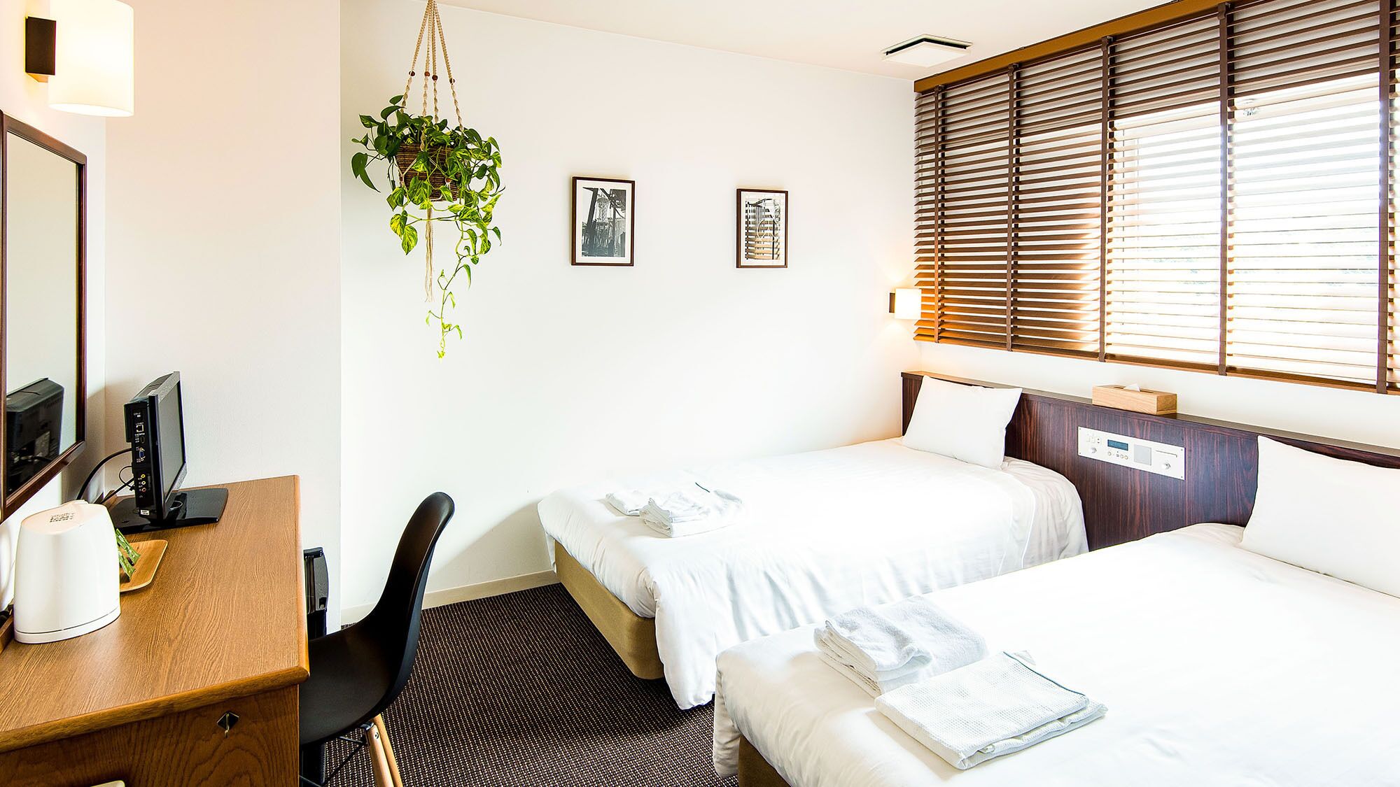 Une chambre d’hôtel avec deux lits, un bureau avec un ordinateur, une chaise, une plante suspendue et des œuvres d’art au mur.