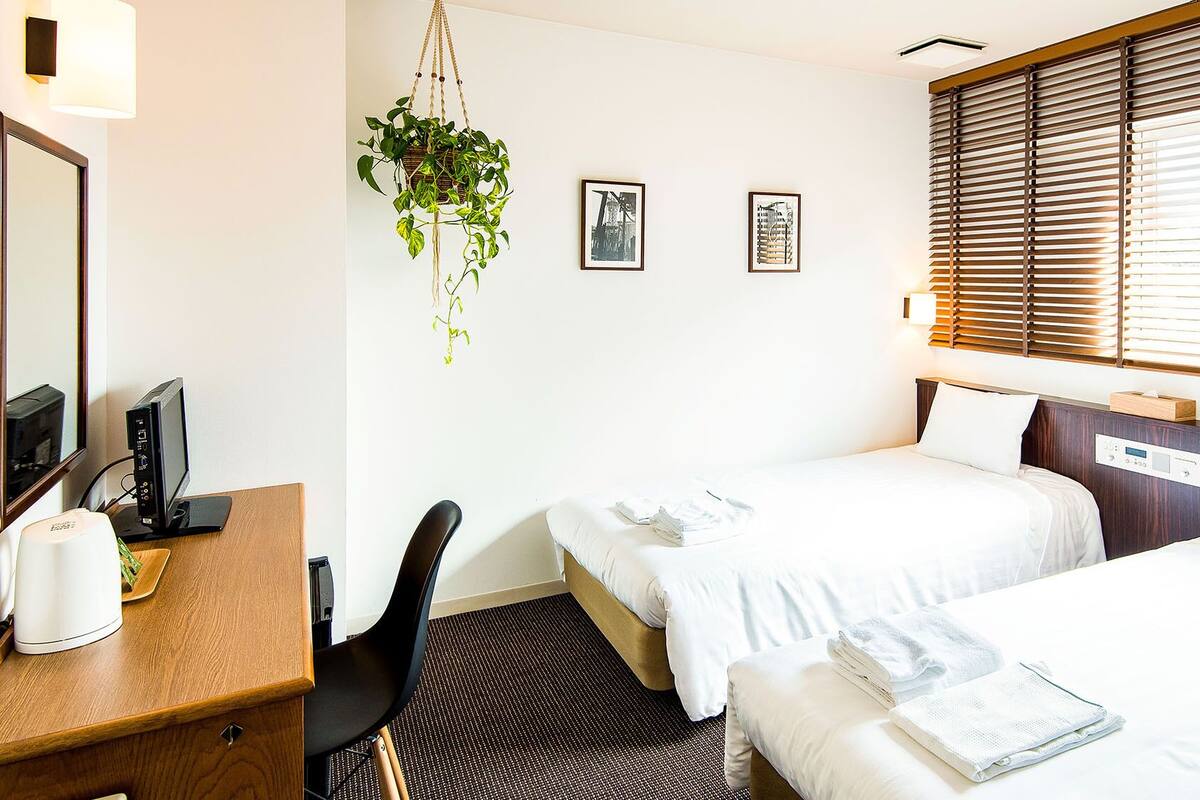 A hotel room with two beds, a desk with a computer, a chair, a hanging plant, and artwork on the wall.