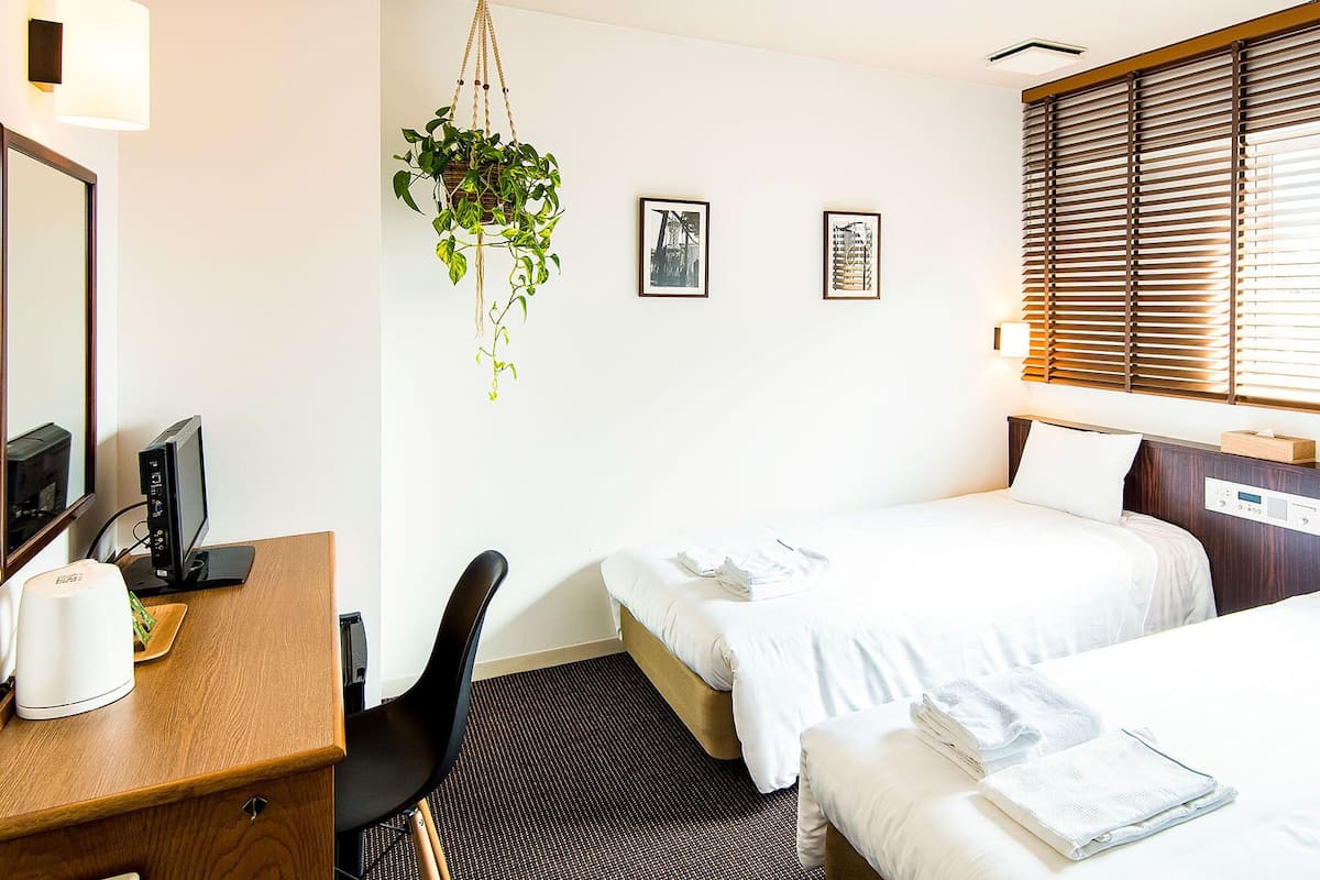 Habitación de hotel con dos camas, un escritorio con computadora, una silla, una planta colgante y obras de arte en la pared.