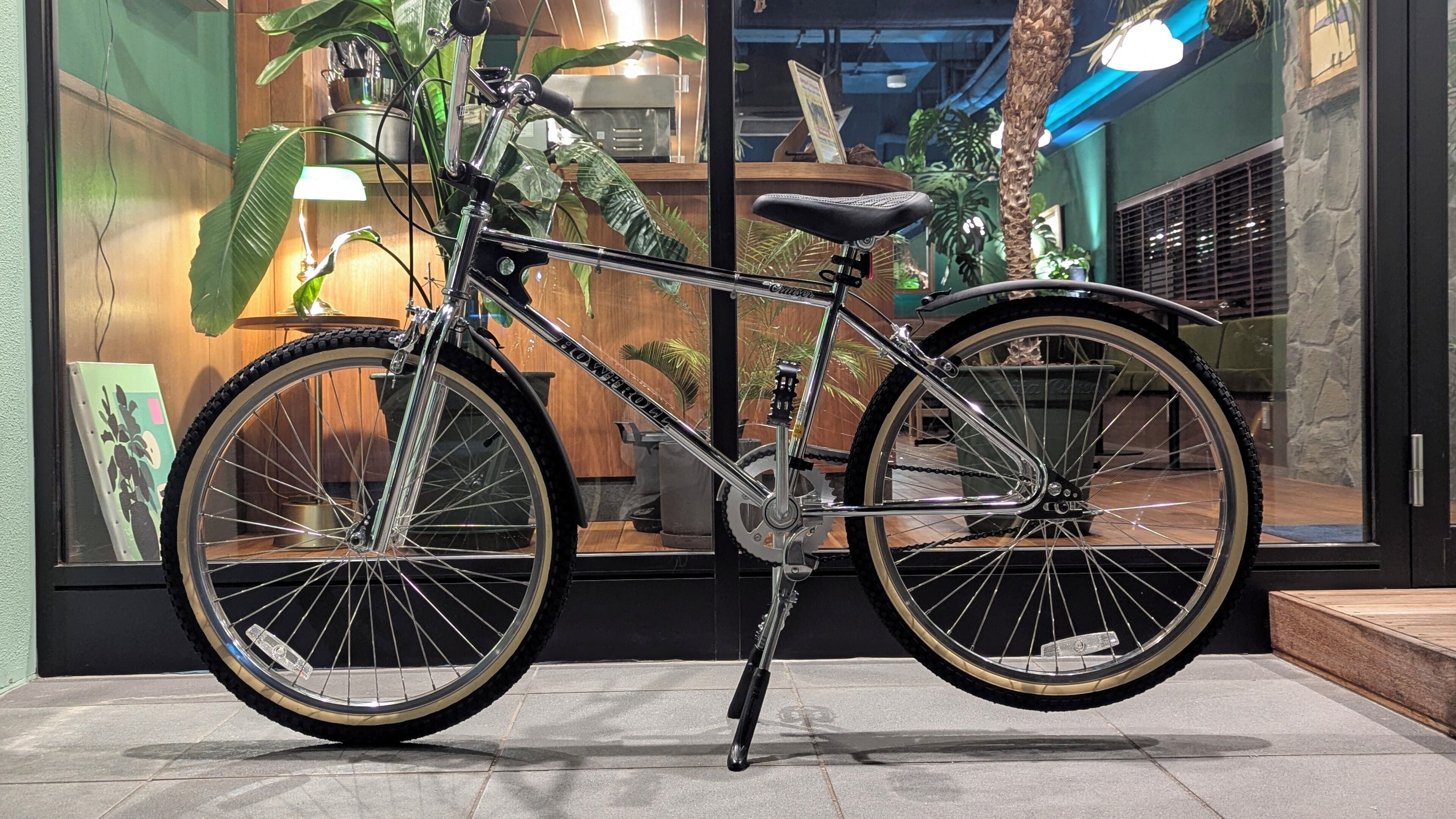En motionscykel med kedja och växlar, placerad inomhus på ett trägolv, med en bakgrund bestående av krukväxter och ett fönster.