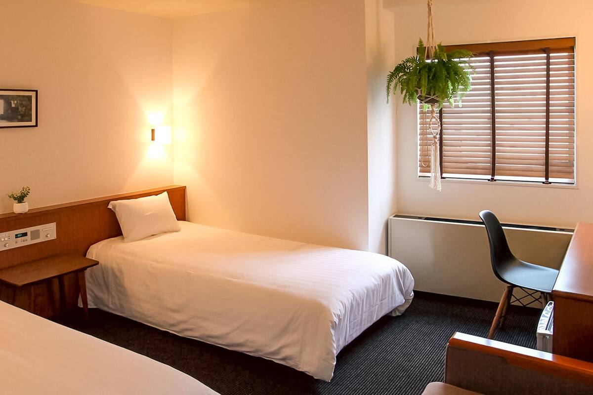 A hotel room with two single beds, a desk with a chair, a small table, a wall-mounted TV, a framed picture, a wall-mounted light, and a window with blinds and a hanging plant.