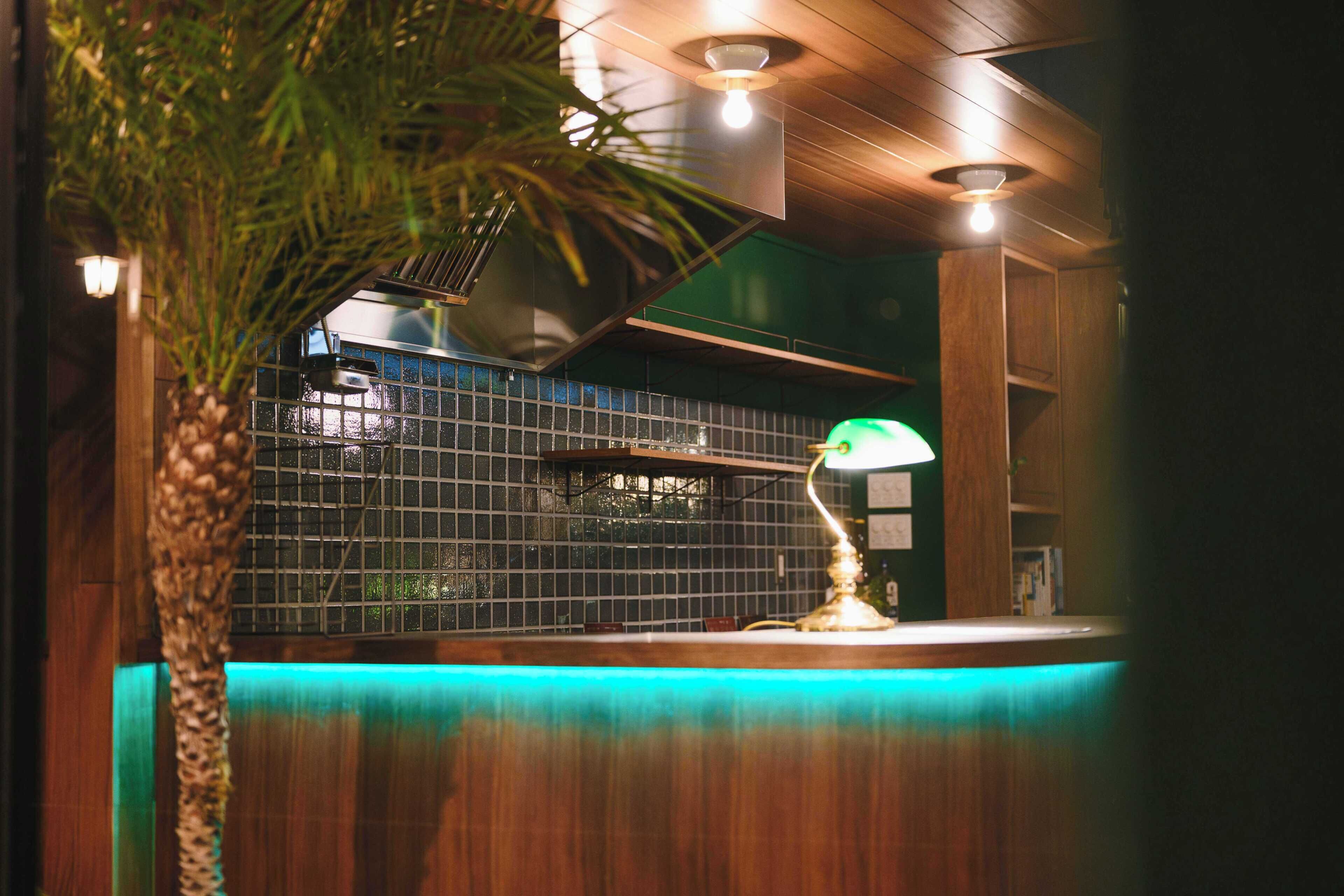 Uma área de bar com balcão iluminado em verde, ventilador de teto e parede com padrão de grade.